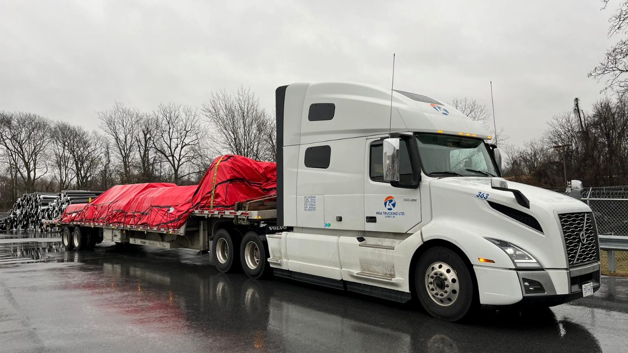 RRA Trucking flatbed unit with tarped freight on a rainy highway, on the road since 2013.