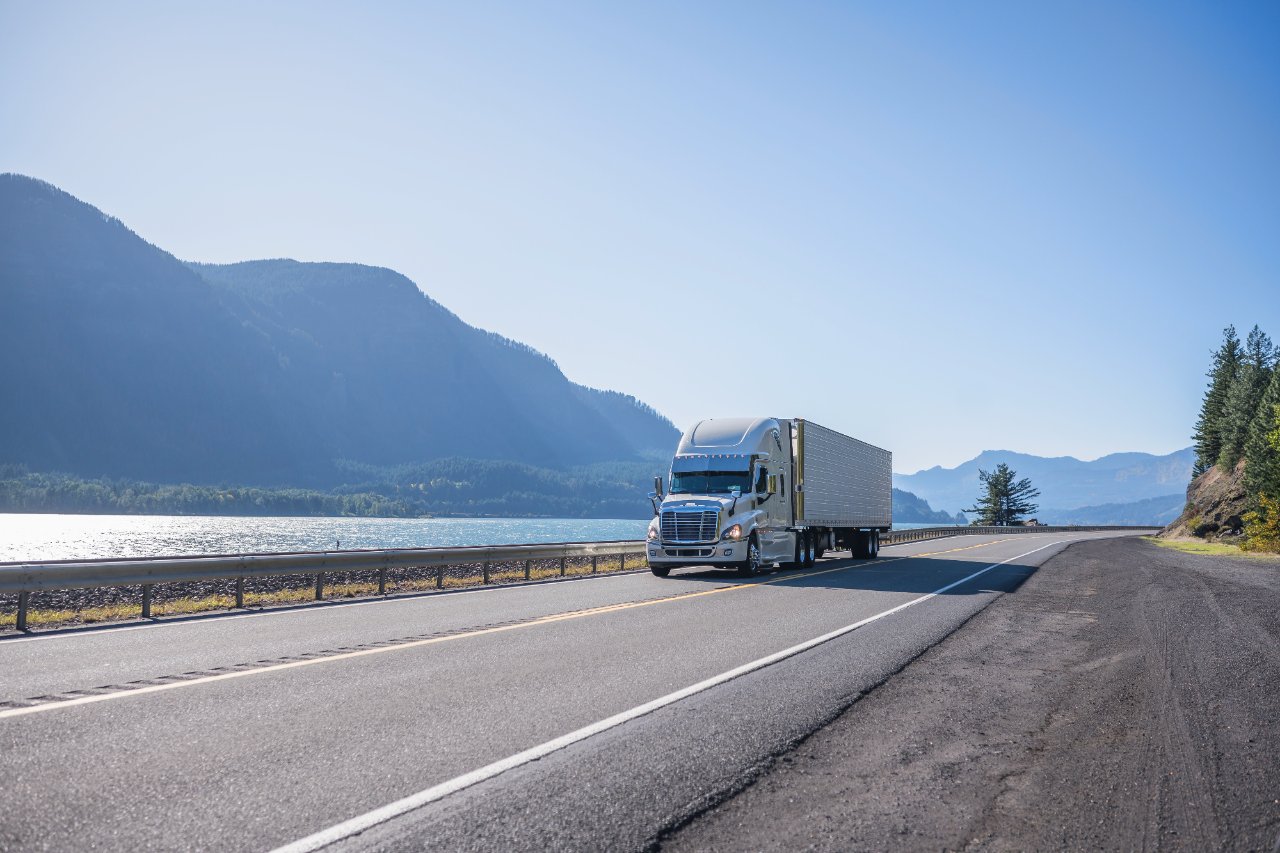 Tractor-trailer on a highway beside a lake and mountains.