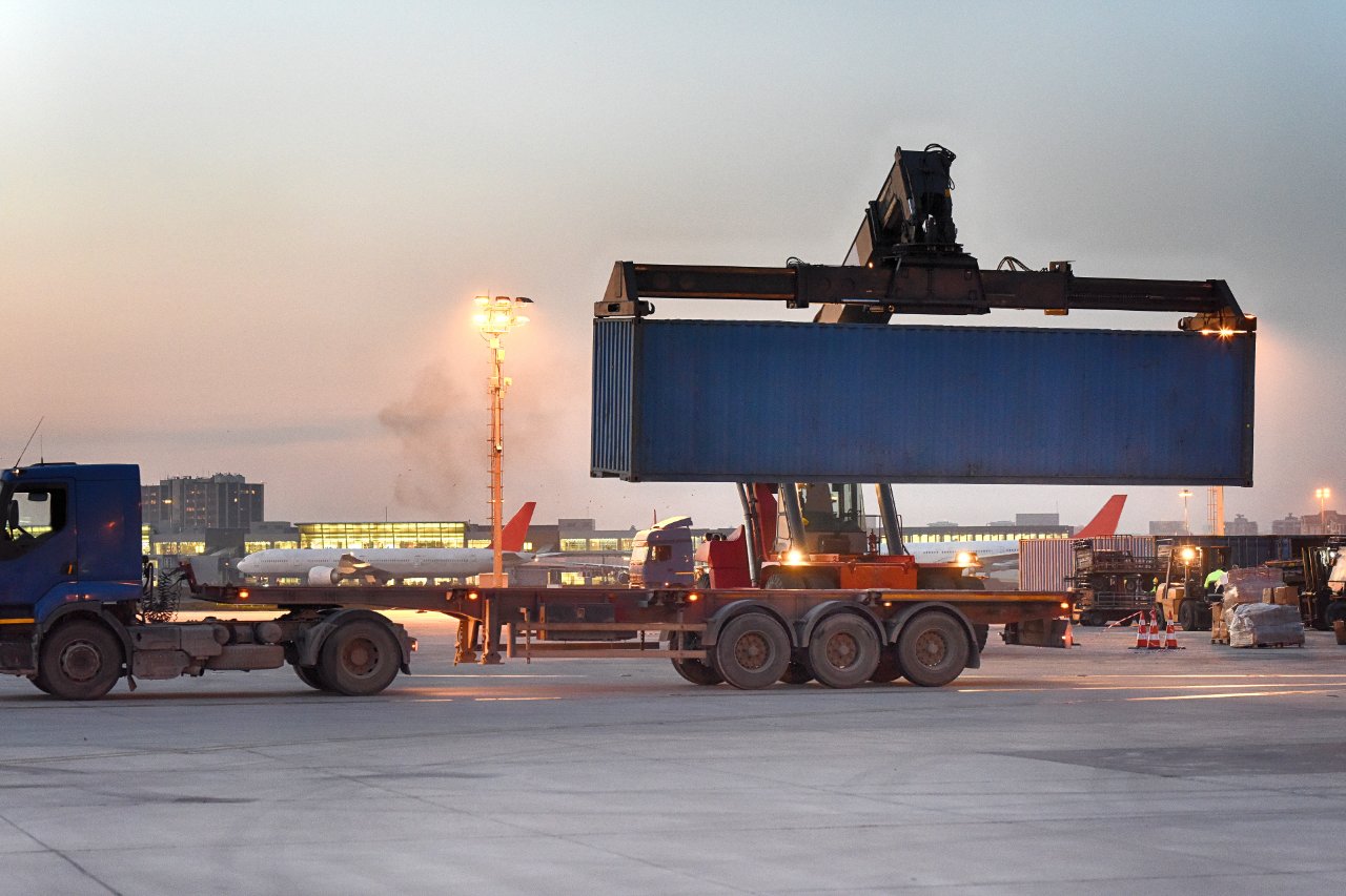 Shipping container being loaded onto a truck chassis at a multimodal freight terminal at dusk.