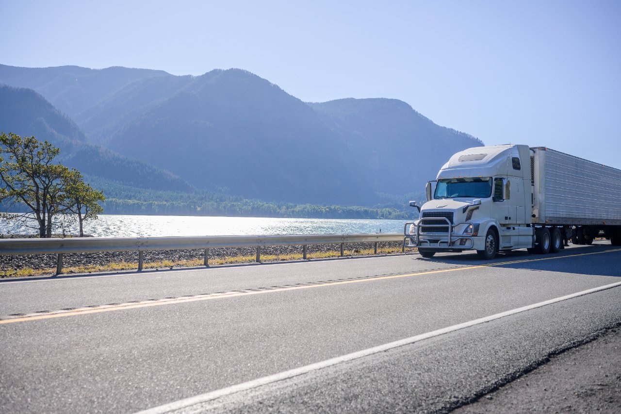 White tractor-trailer on a highway beside a lake and mountains.