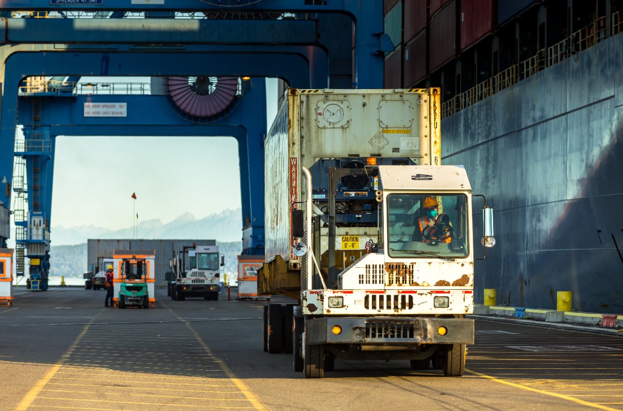 Terminal tractor hauling a shipping container along the berth at a container port, mountains in the distance.