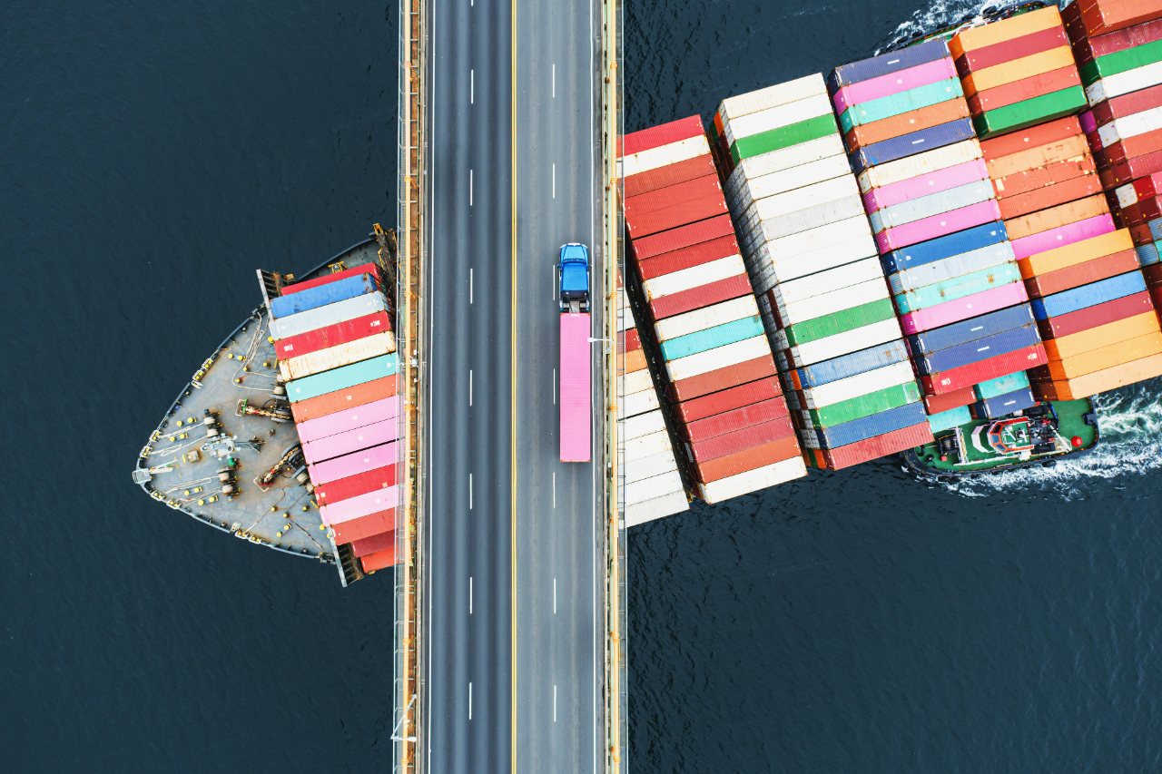 Aerial view of a container ship, a truck crossing a bridge, and stacked shipping containers — ocean and road freight in one connected chain
