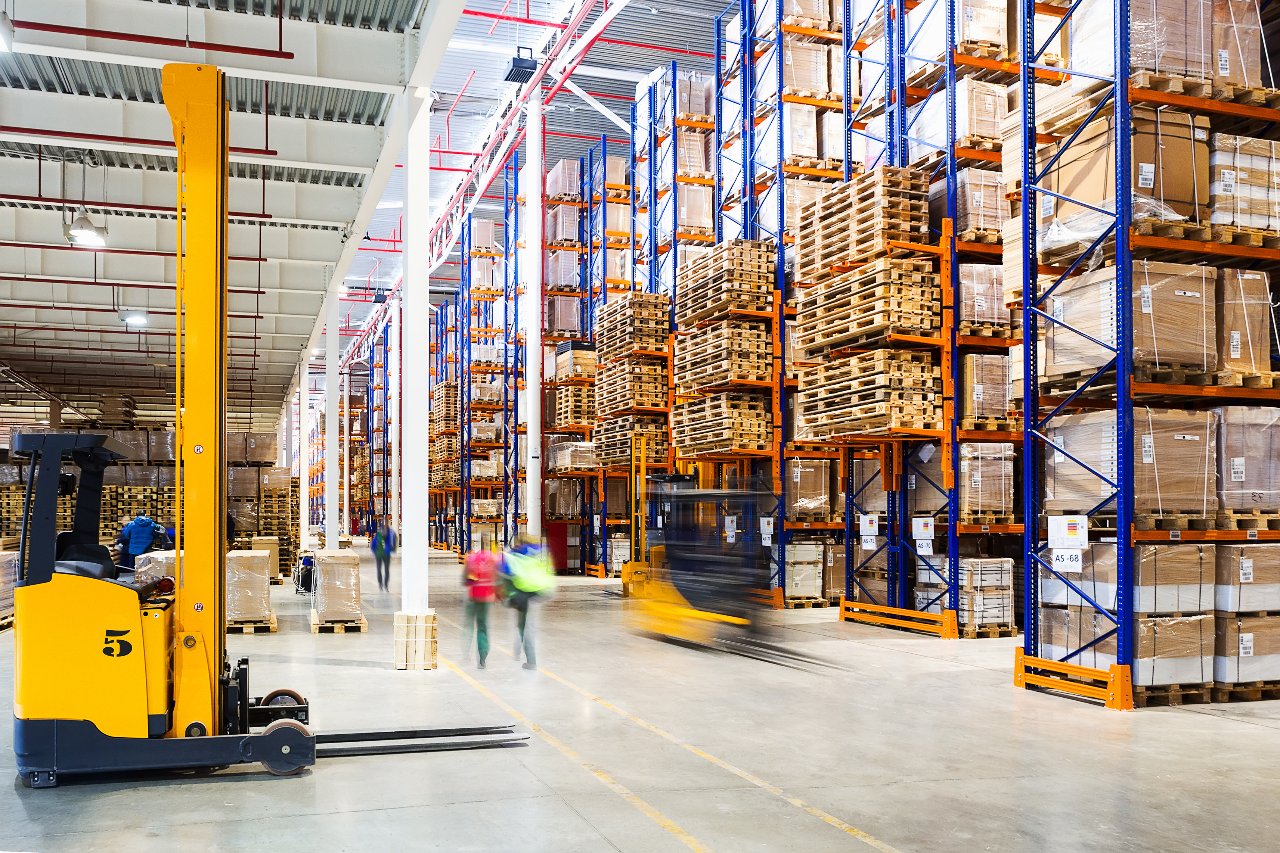 Yellow reach truck in a high-bay racking warehouse with pallets and moving operators.